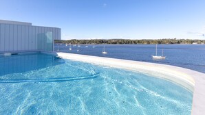 A swimming pool with clear blue water, surrounded by a white pool deck, and a view of the sea with sailboats in the distance. - Joy at Ohana: Balcony & Pool Overlooking the Bay (Batemans Bay)