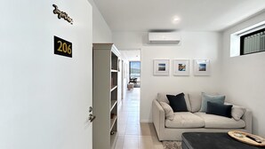 A modern living room with a beige sofa, a dark ottoman, and a small coffee table. There is a door with a number sign, a hallway leading to a balcony, and a mounted air conditioner. - Joy at Ohana: Balcony & Pool Overlooking the Bay (Batemans Bay)