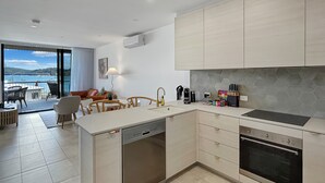 A modern kitchen with a dishwasher, oven, and a dining area with a view of the sea. - Joy at Ohana: Balcony & Pool Overlooking the Bay (Batemans Bay)