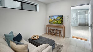 A modern living room with a sofa, a coffee table, and a flat-screen TV on a stand. - Joy at Ohana: Balcony & Pool Overlooking the Bay (Batemans Bay)