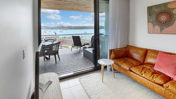 A modern living room with a leather sofa, a small white table, and a balcony with a view of the sea. - Joy at Ohana: Balcony & Pool Overlooking the Bay (Batemans Bay)