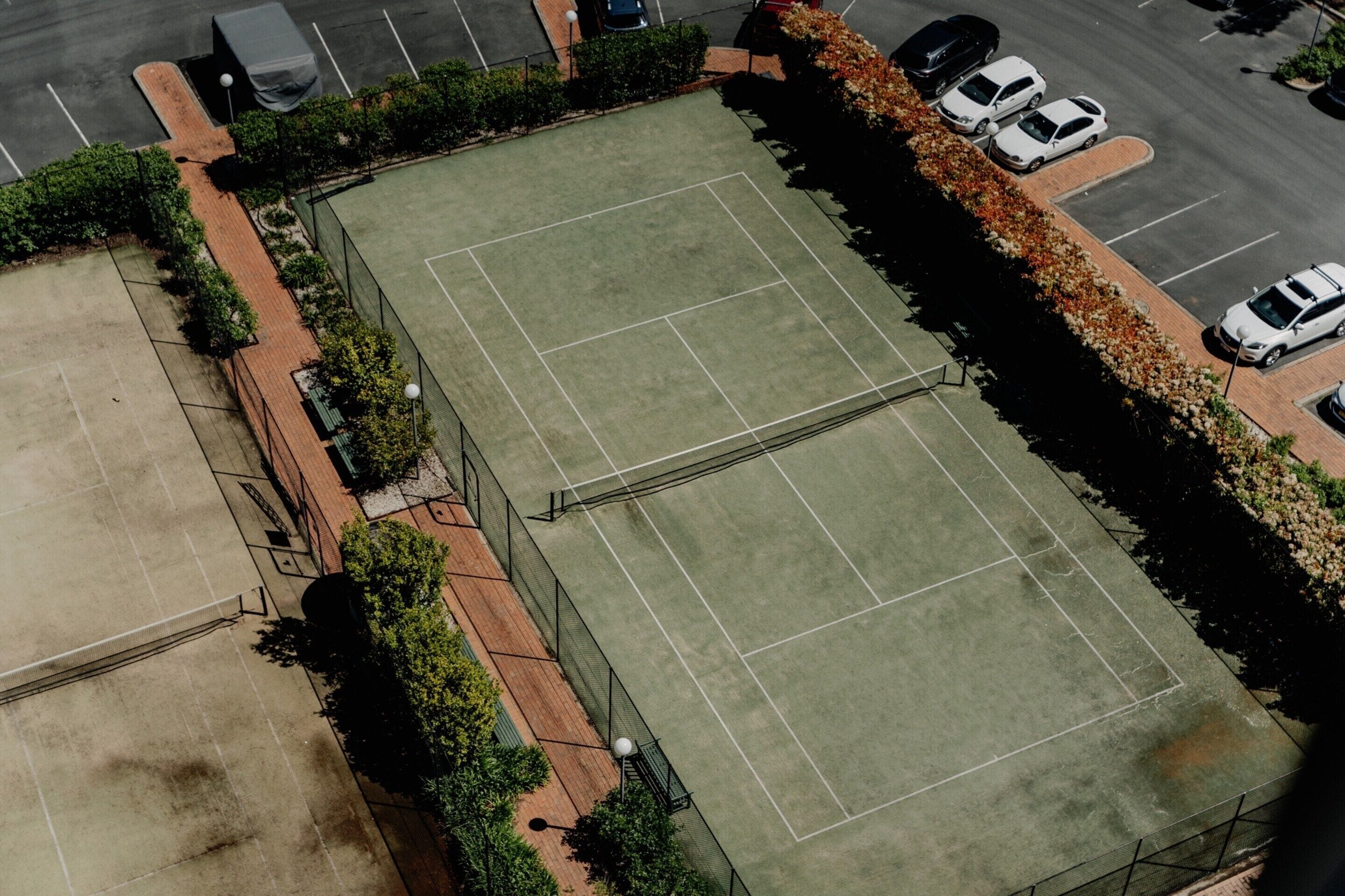 An aerial view of a tennis court complex with multiple courts, a parking area, and surrounding landscaping.
