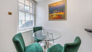 A dining area with a glass table and green upholstered chairs, a painting on the wall, and a window with a view of buildings.