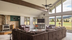 A spacious living room with a large sectional sofa, a stone fireplace, leather armchairs, and a television mounted on the wall. - Fairway Views | Ski Trail | Hot Tub (Big Sky)
