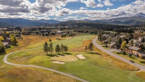 An aerial view of a golf course with a winding road, residential buildings, and a mountainous backdrop. - Fairway Views | Ski Trail | Hot Tub (Big Sky)
