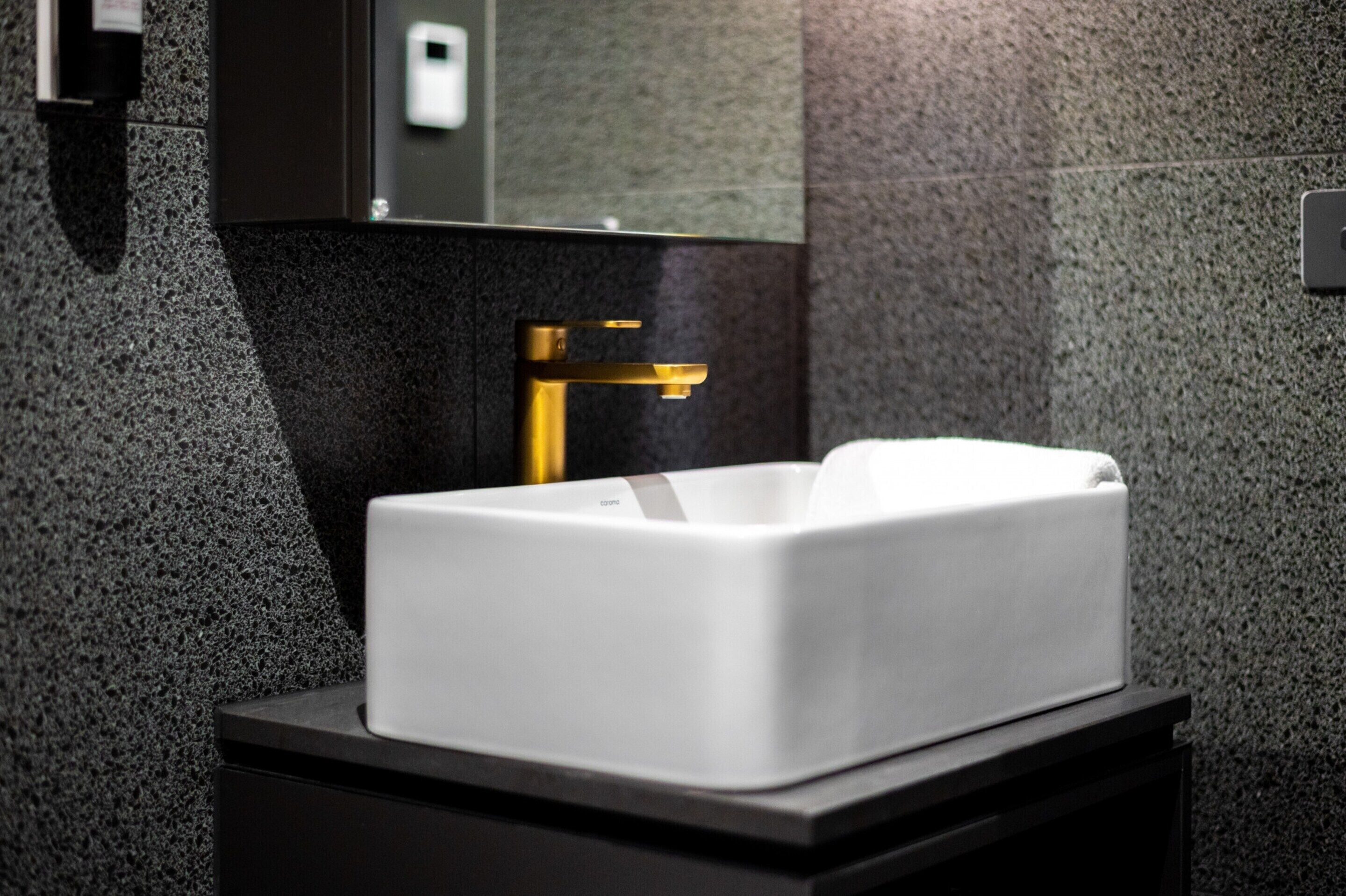 A modern bathroom with a white sink, a gold faucet, and a dark granite countertop.
