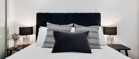Una cama bien hecha con cabecera negra, dos mesitas de noche con lámparas y almohadas estampadas.