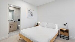 Un dormitori modern amb un llit de fusta, una taula de nit amb una làmpada, una petita taula auxiliar i una obra d'art emmarcada a la paret, amb un bany visible a través d'una porta oberta.
