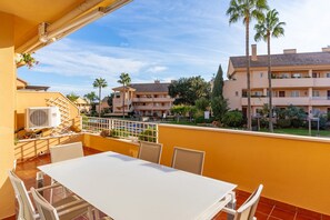 Un balcon avec une table et des chaises blanches, donnant sur un quartier résidentiel agrémenté de palmiers et d’une piscine.