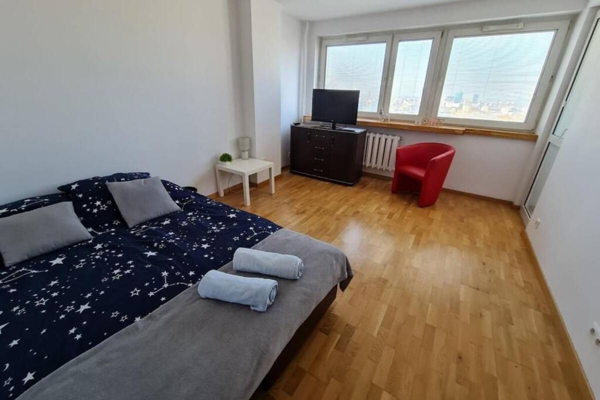 Ein Schlafzimmer mit einem Bett, einem Fernseher, einem roten Sessel und einem Fenster mit Blick auf die Stadt.