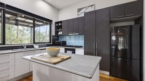 A modern kitchen with dark cabinets, a stainless steel refrigerator, and a large window overlooking a forest. - Stylish 3-bedroom and Outdoor Grill and Pizza Oven (Drysdale)