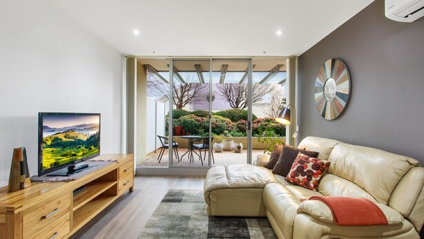 A modern living room with a television, leather sofa, and a view of a garden through glass doors. - Madecomfy Spacious Canberra LI (Canberra)