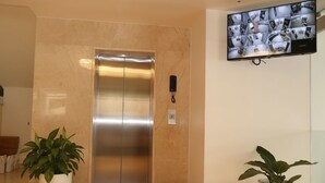 Un ascensor moderno con panel de control, cámara de seguridad en la pared y plantas en macetas en un área de recepción.