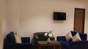 Una sala de estar con dos sofás azules, una pequeña mesa de centro de madera, un jarrón con flores y un televisor montado en la pared.
