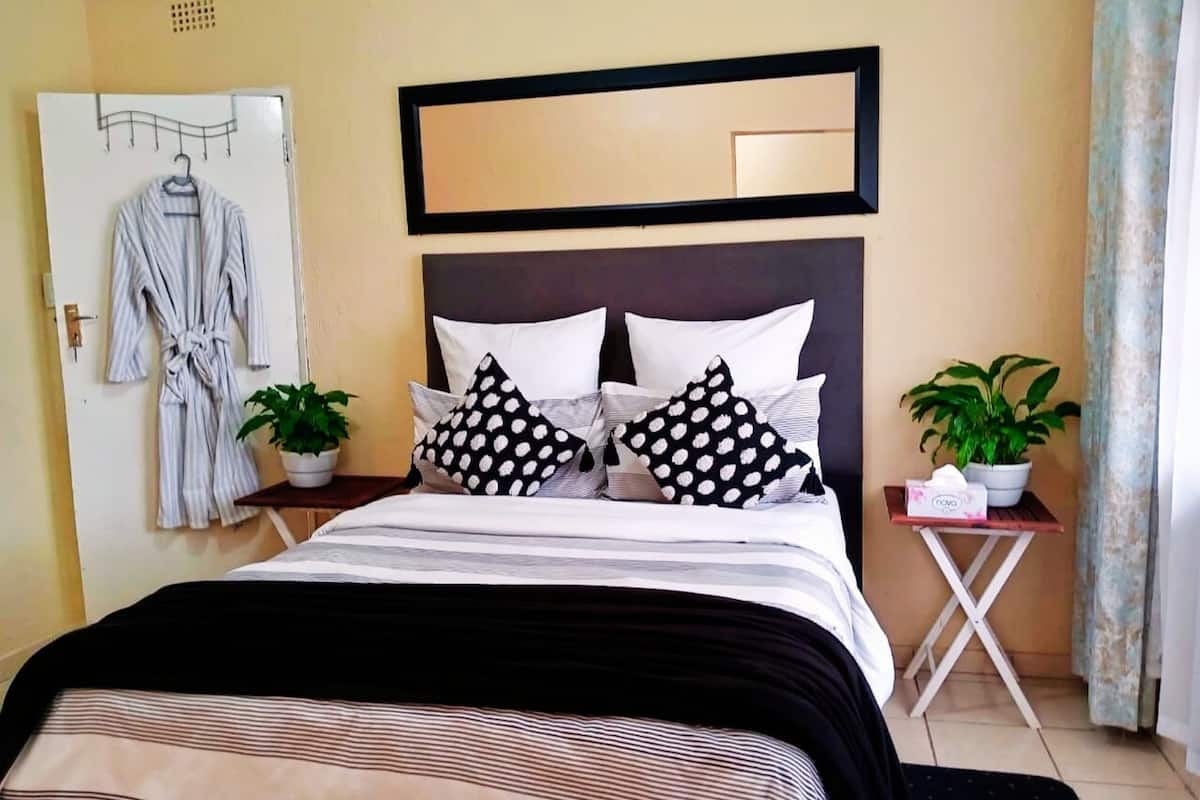 Un lit bien fait, avec une literie à rayures, deux plantes en pot, un miroir au-dessus de la tête de lit et un peignoir accroché à une porte.
