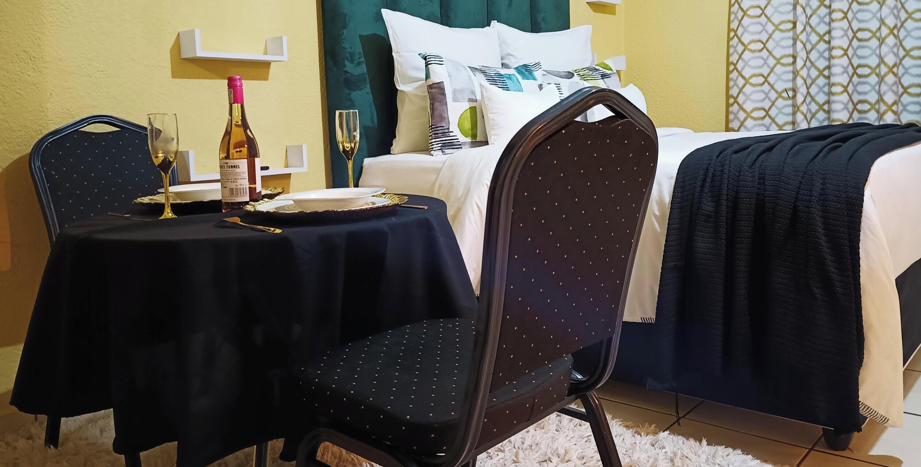 A hotel room with a dining table set for two, a bed with a dark blue bedspread, and a window with patterned curtains.