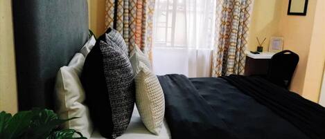 A hotel room with a bed, pillows, a bedside table with a lamp, and a window with curtains.