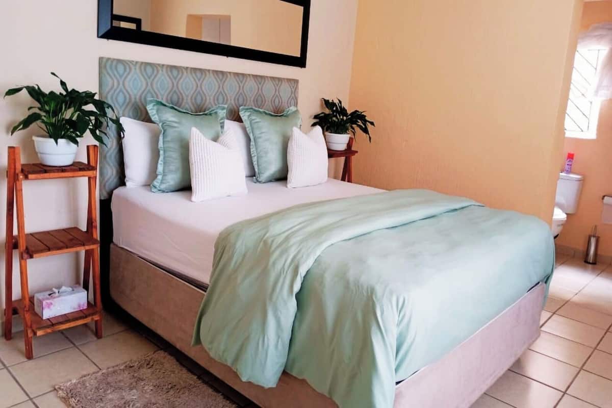 Une chambre à coucher bien rangée, avec un lit, des oreillers, une table de chevet, une étagère avec des plantes et un miroir au mur.