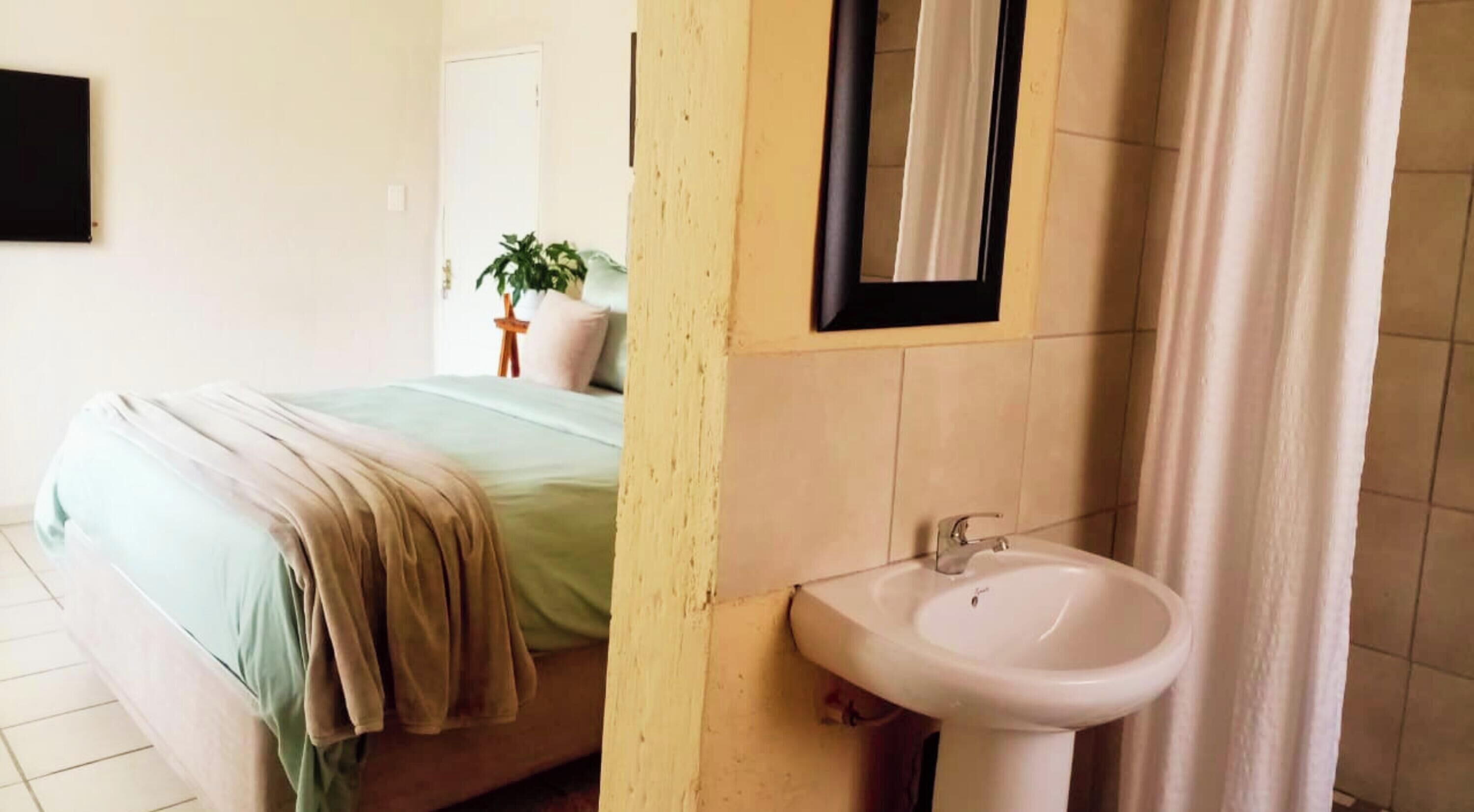 A bedroom with a bed, a bedside table, a mirror, and a bathroom with a sink and a shower curtain.