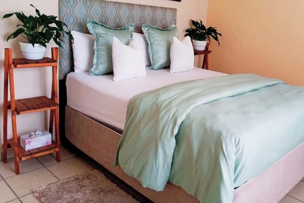A bed with a light green bedspread, a wooden nightstand with a potted plant, and a headboard with a geometric pattern.