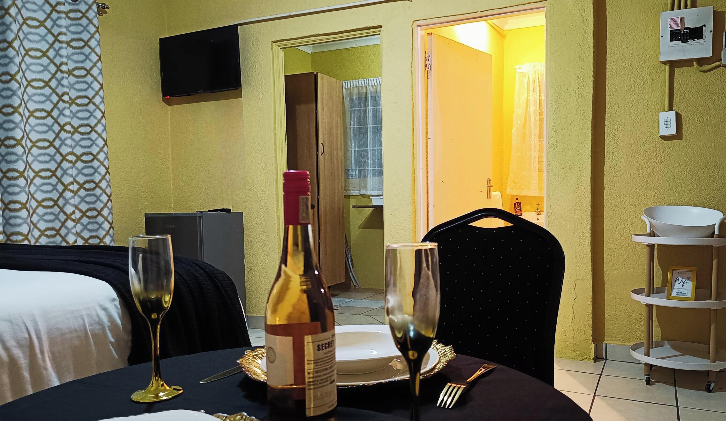 A hotel room with a bed, a dining table with wine glasses and a bottle, a TV mounted on the wall, and a bathroom visible through an open door.