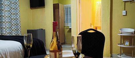 A hotel room with a bed, a dining table with wine glasses and a bottle, a TV mounted on the wall, and a bathroom visible through an open door.