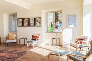 A spacious living room with a large window, multiple seating options, and wall-mounted artwork.