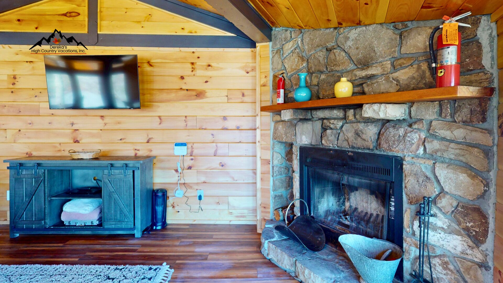Interior acogedor de una cabaña con chimenea de piedra, paredes de madera, televisor montado sobre la chimenea y un banco rústico de madera con una cesta.