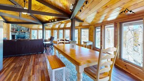 A spacious wooden cabin with a large dining table, a fireplace, and large windows offering views of the surrounding forest.