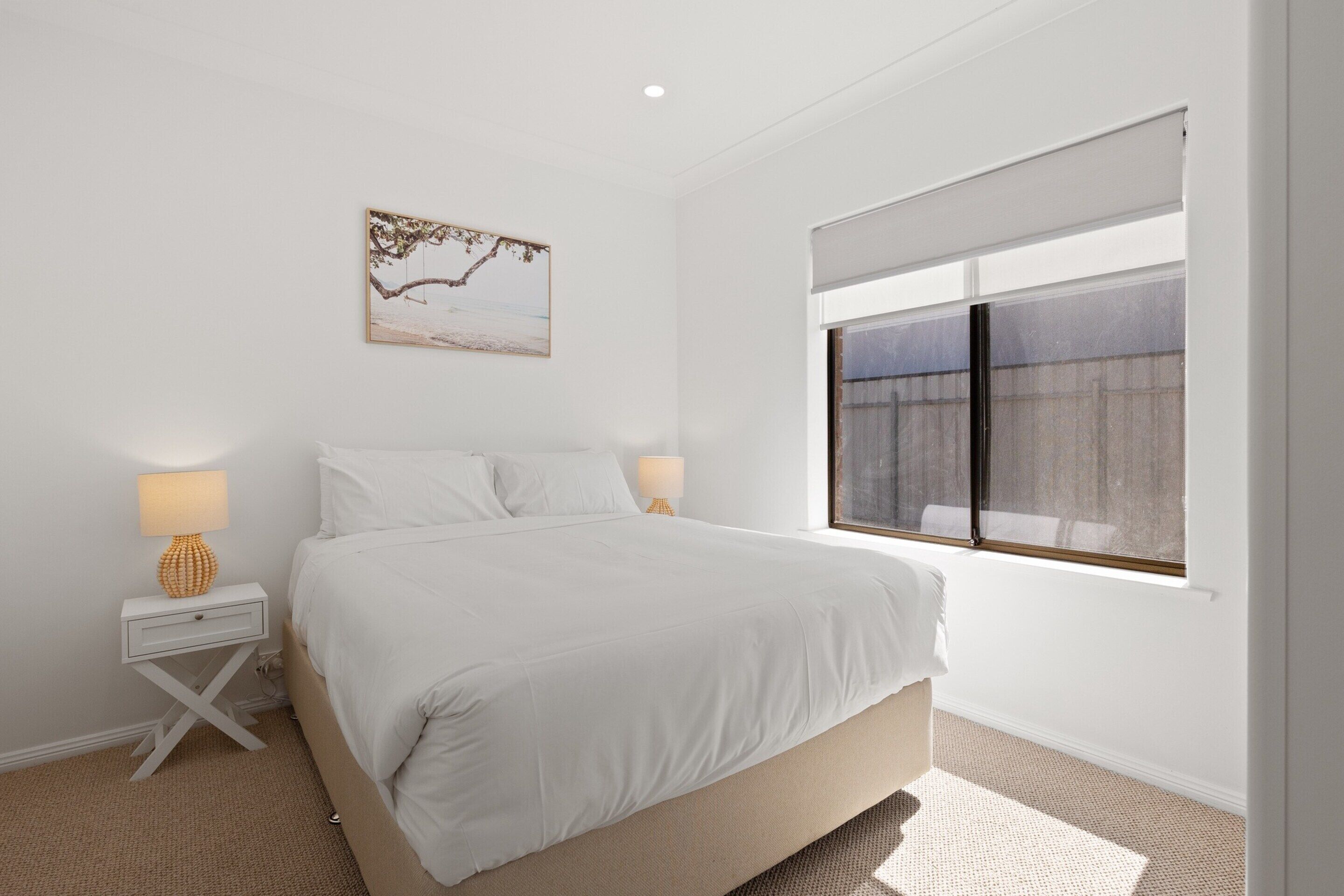 A bedroom with a bed, two bedside tables with lamps, a window with blinds, and a wall-mounted picture.