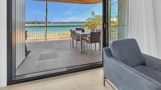 A balcony with a table and chairs, a grey sofa, and a view of the sea. - 3-bed: Riverfront Alfresco Dining & Rooftop Pool (Batemans Bay)