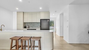A modern kitchen with a central island, bar stools, and a refrigerator. - 3-bed: Riverfront Alfresco Dining & Rooftop Pool (Batemans Bay)