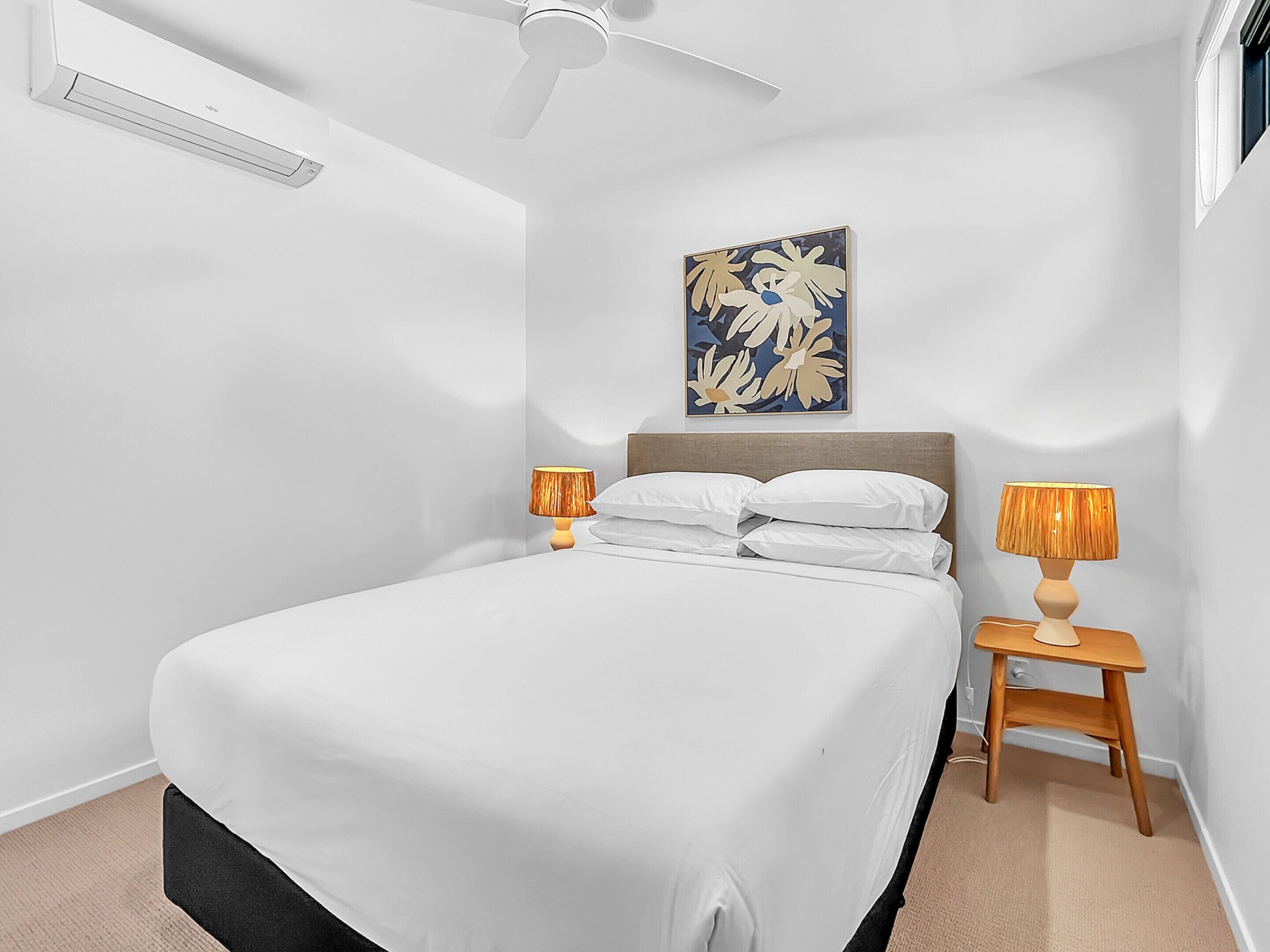 Un dormitorio con una cama, dos mesitas de noche, un ventilador de techo, aire acondicionado y una obra de arte colgada en la pared.