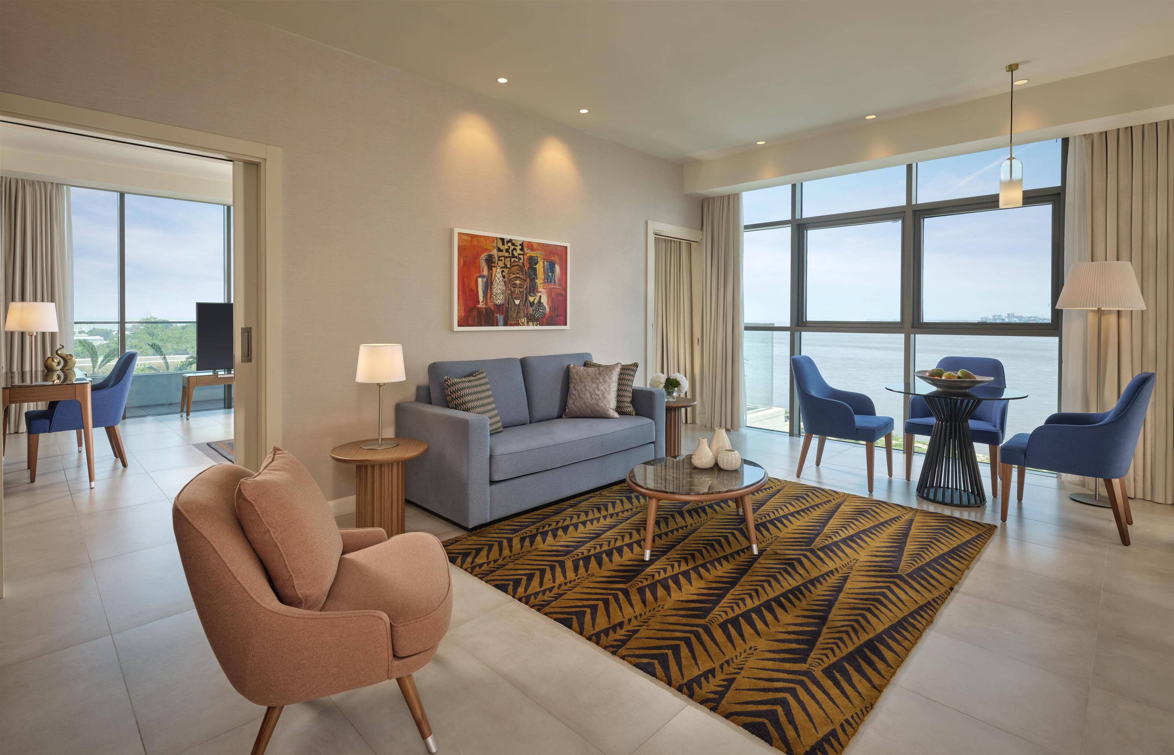 A modern hotel room with a sofa, armchair, dining table, and chairs.