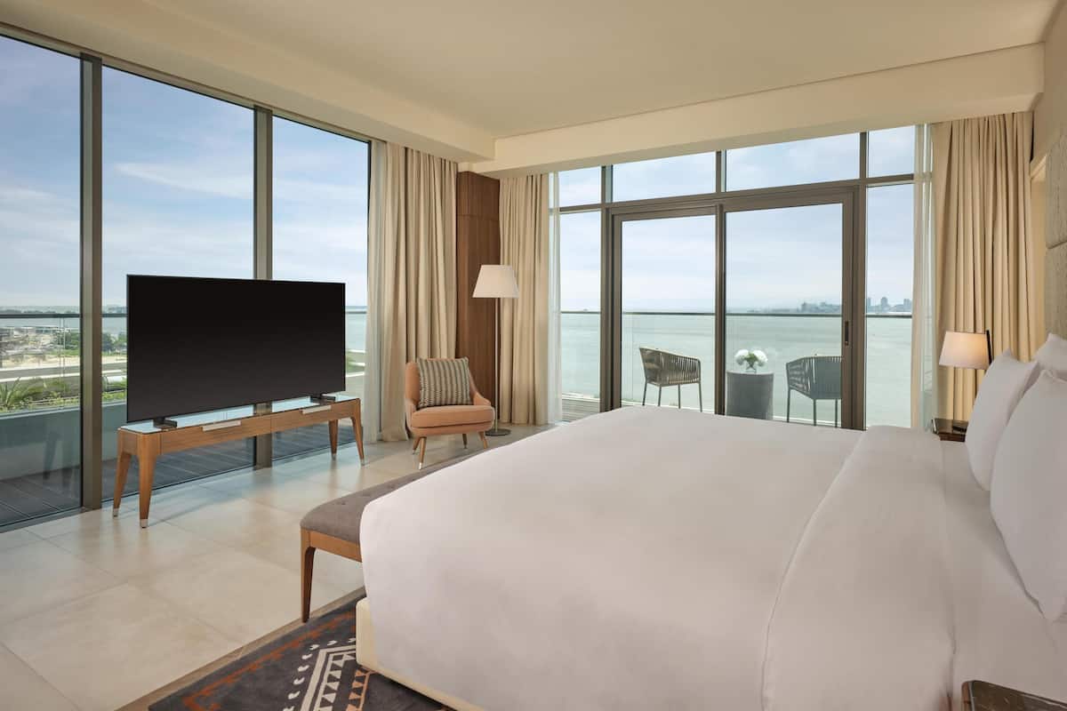 A hotel room with a large bed, a TV on a stand, a chair, a small table, and a view of the water.