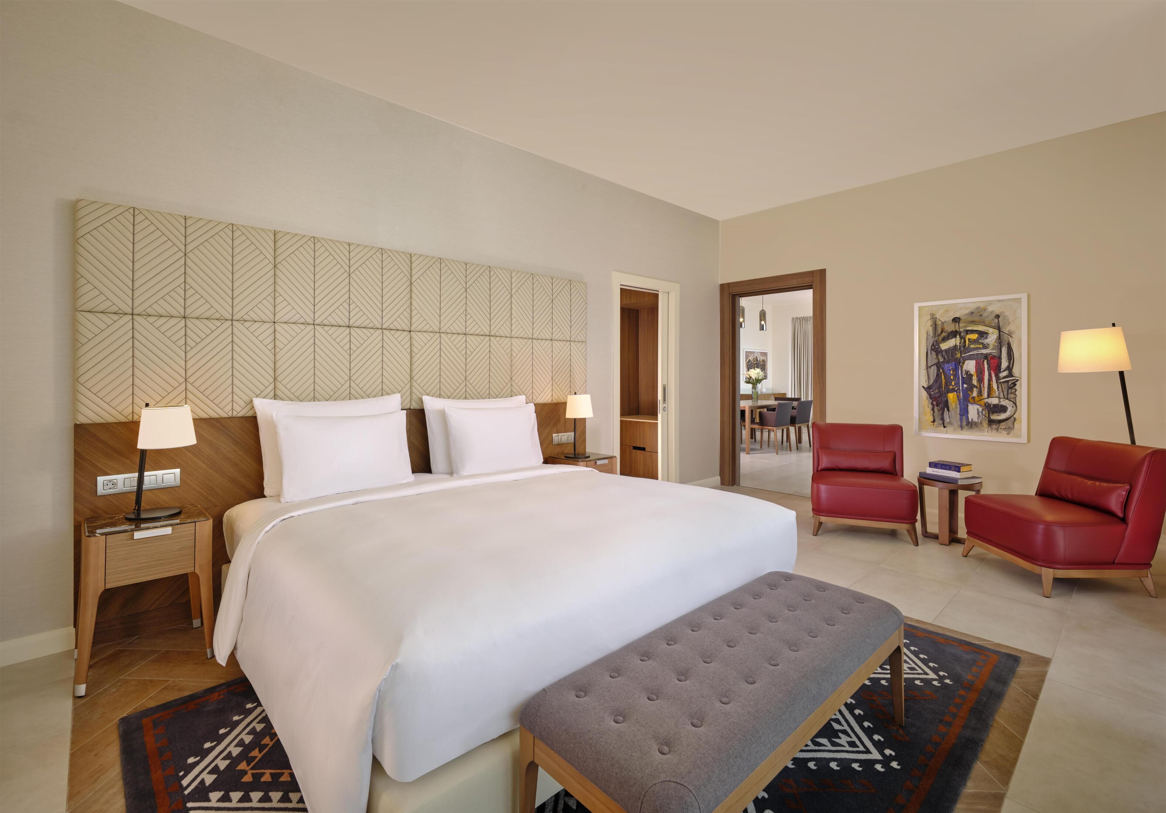 Habitación de hotel con una cama grande, dos mesitas de noche, un banco, un sillón rojo, una mesita pequeña y un cuadro en la pared.