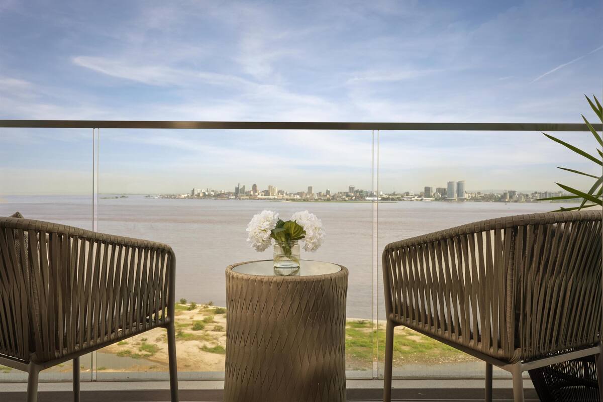 A balcony with two wicker chairs, a round table with a vase of flowers, and a view of a city skyline across a body of water.