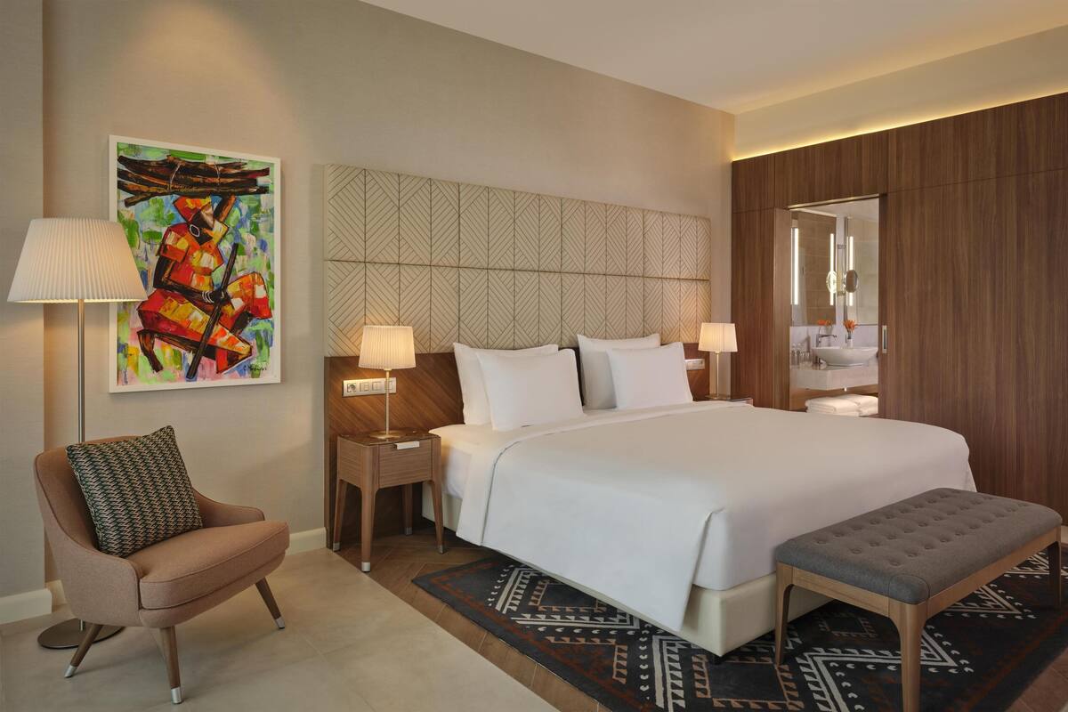 A hotel room with a large bed, a chair, a bedside table, a lamp, a painting on the wall, and a patterned rug.