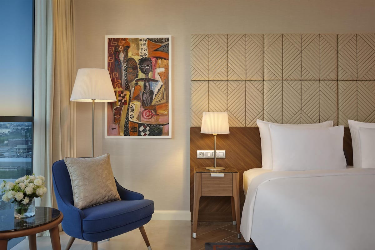 A hotel room with a bed, a blue armchair, a small table with a vase of flowers, a wall-mounted painting, and a geometric-patterned headboard.