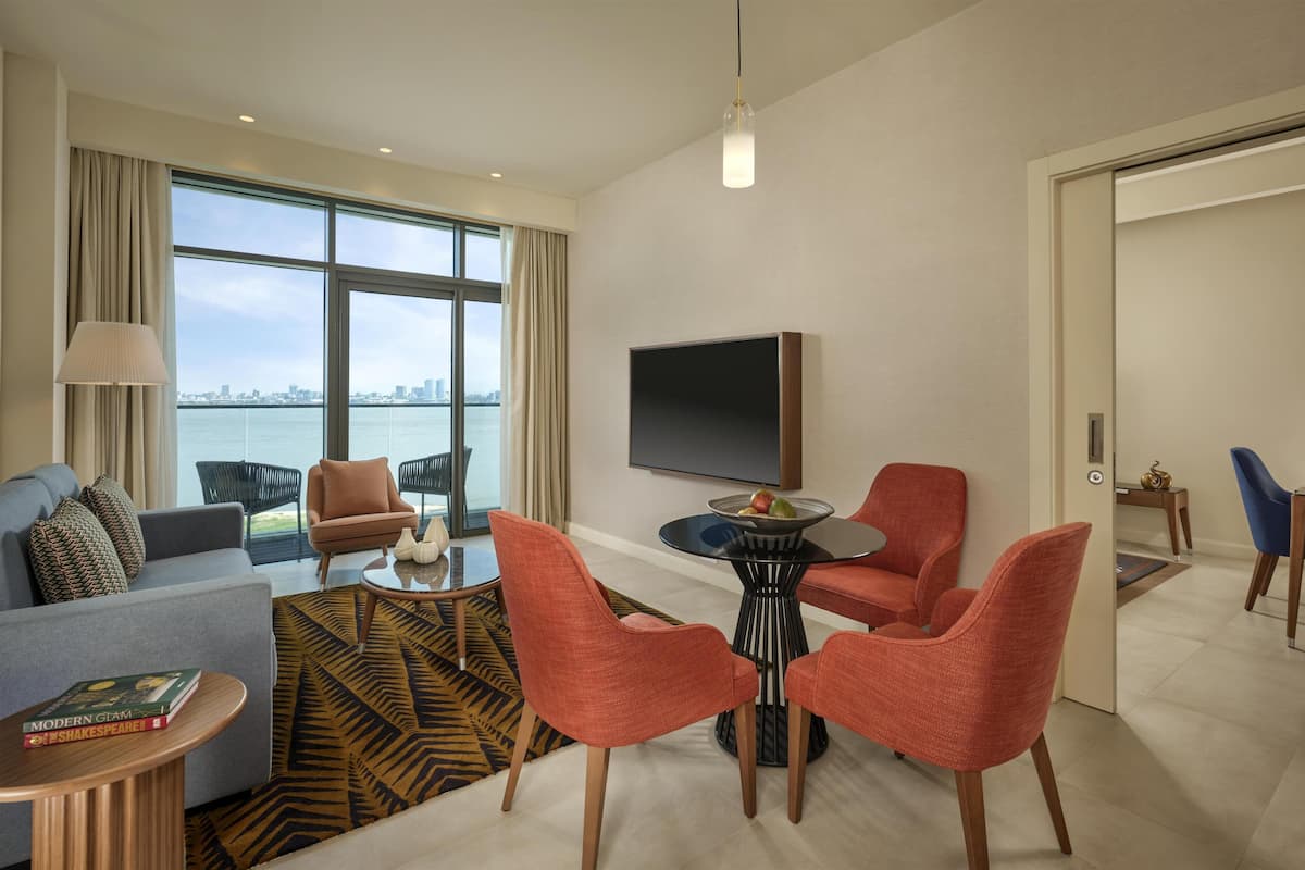 A modern hotel room with a large window, a flat-screen TV, a sofa, a coffee table, and a dining area with chairs.