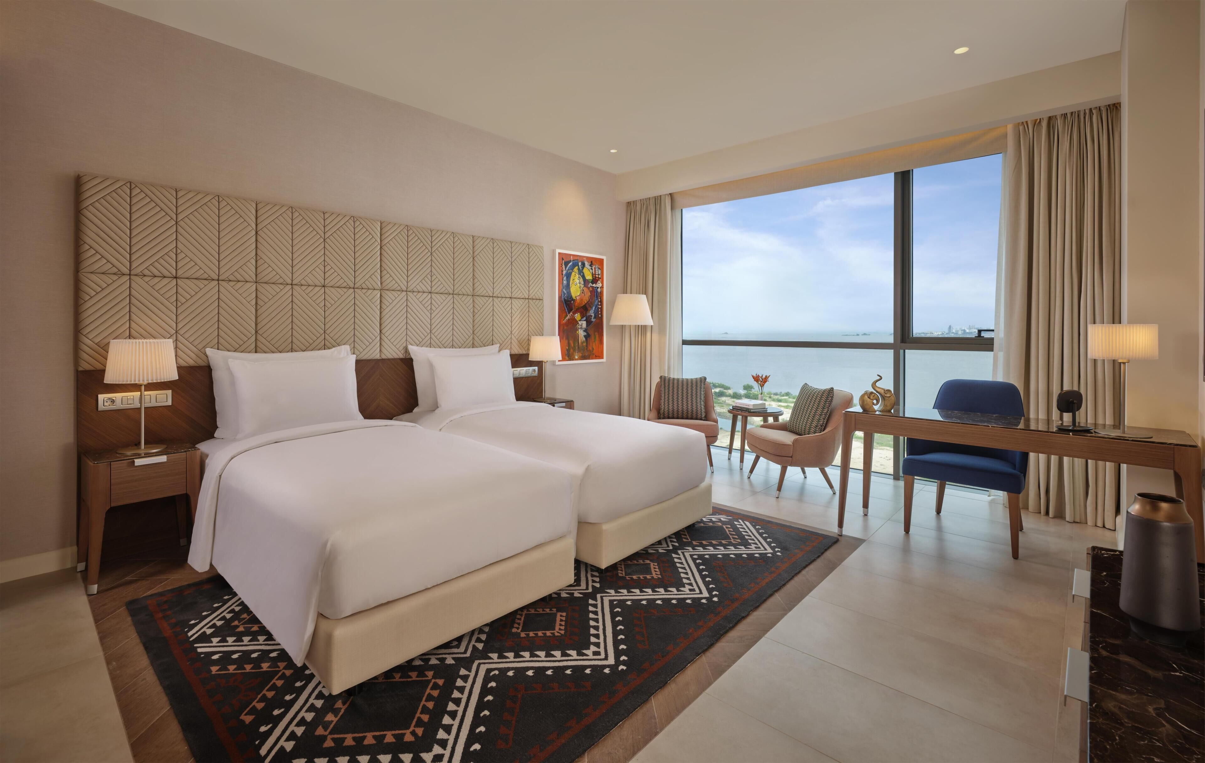 Ein Hotelzimmer mit zwei Betten, einem Schreibtisch, einem Stuhl und Blick auf das Meer, sowie einem gemusterten Kopfteil.