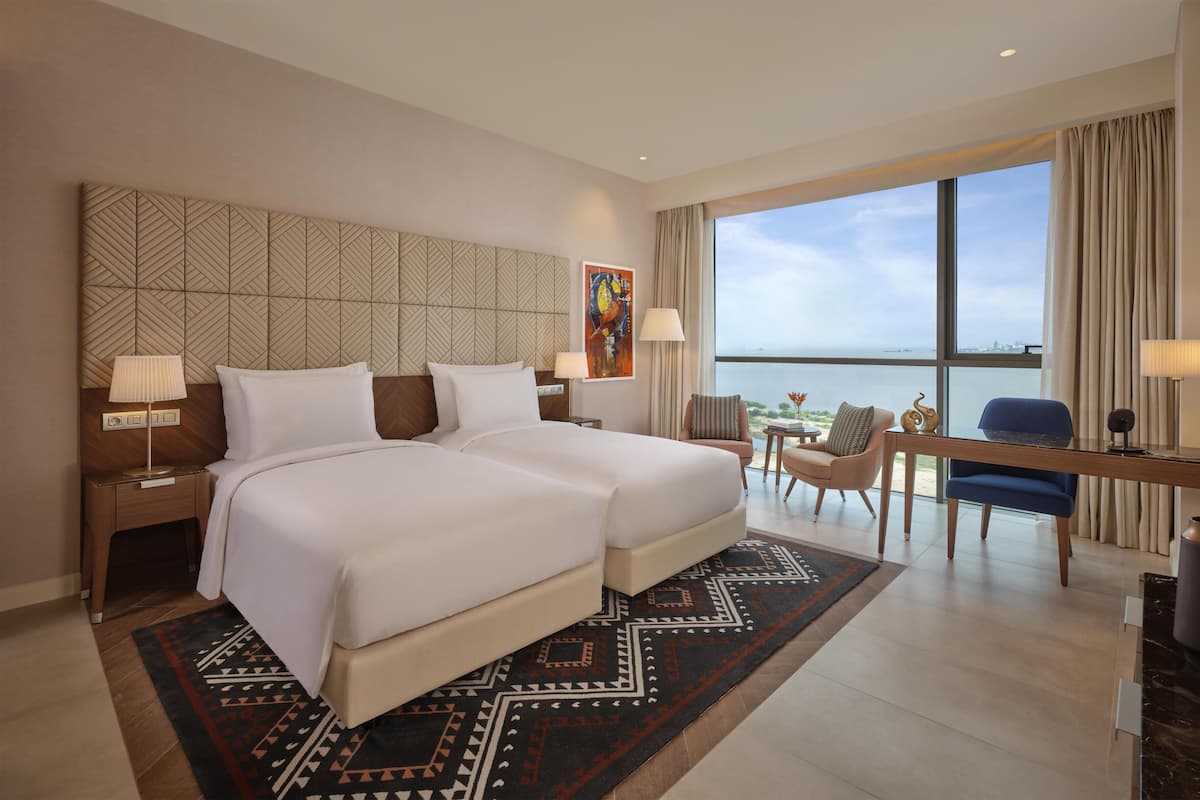 A hotel room with two beds, a desk, a chair, a view of the sea, and a patterned headboard.