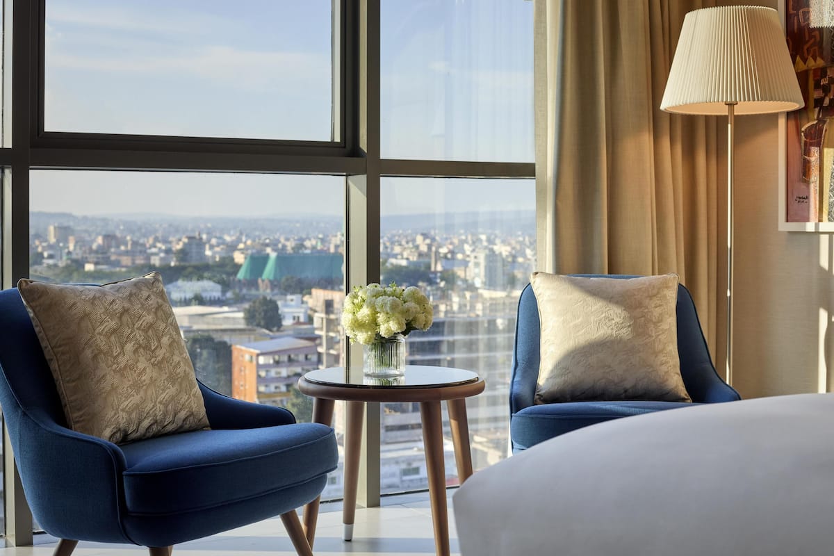A hotel room with a large window offering a city view, two blue armchairs with decorative pillows, a small round table with a vase of flowers, and a floor lamp.