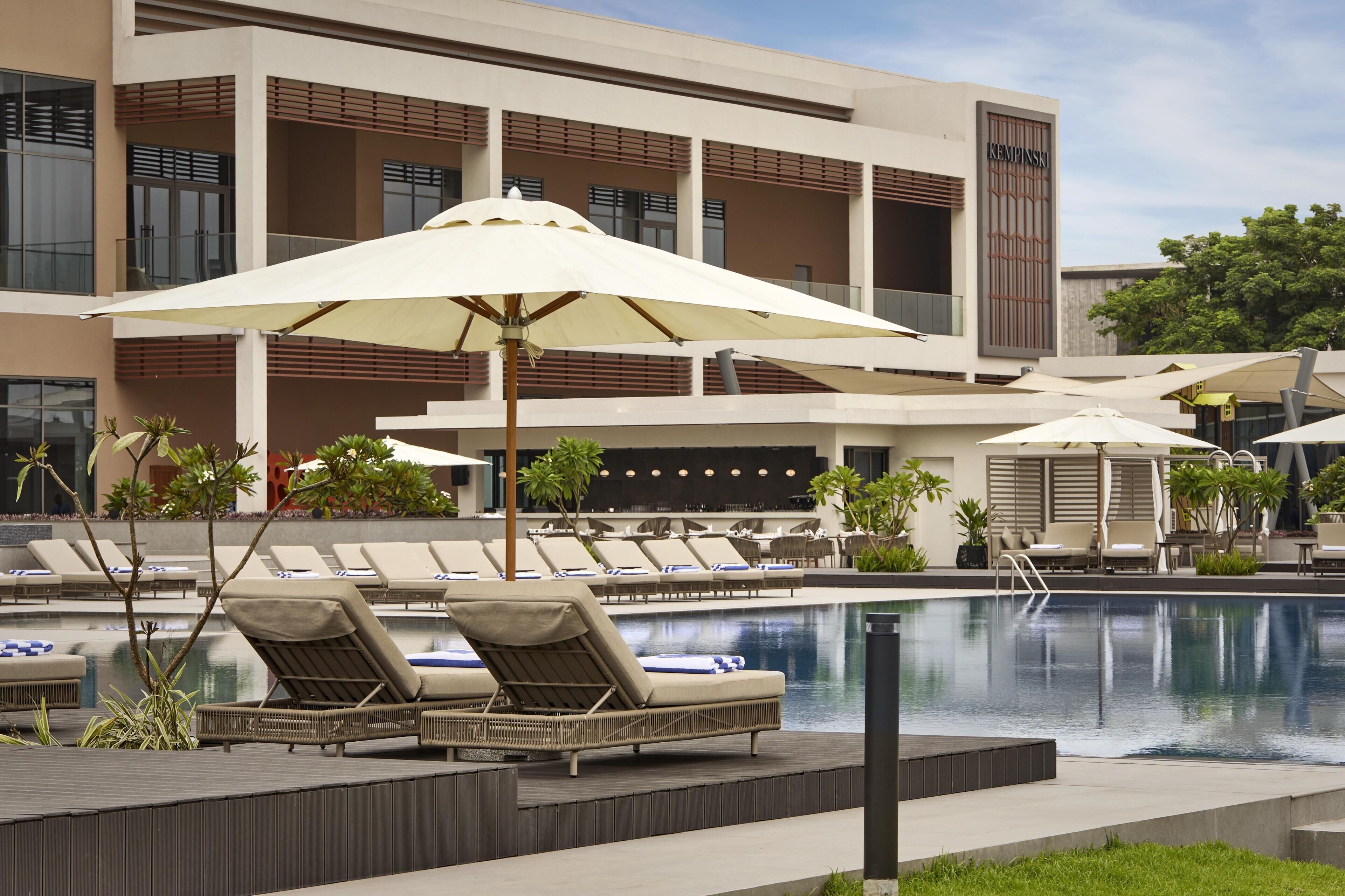 A poolside area with lounge chairs and umbrellas, a modern building with balconies, and a clear sky.