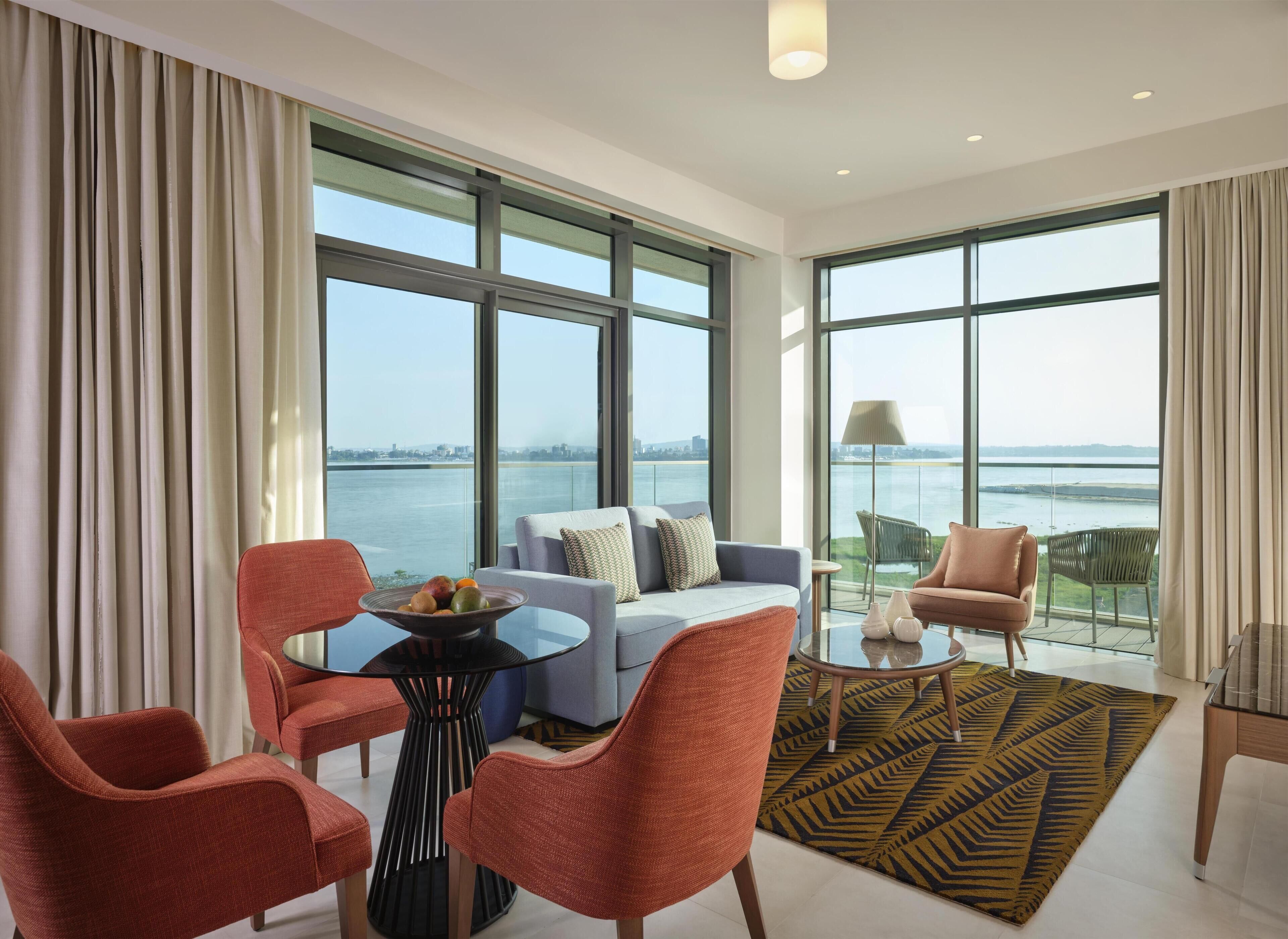A modern living room with large windows, a sofa, two armchairs, a glass coffee table, and a patterned rug.