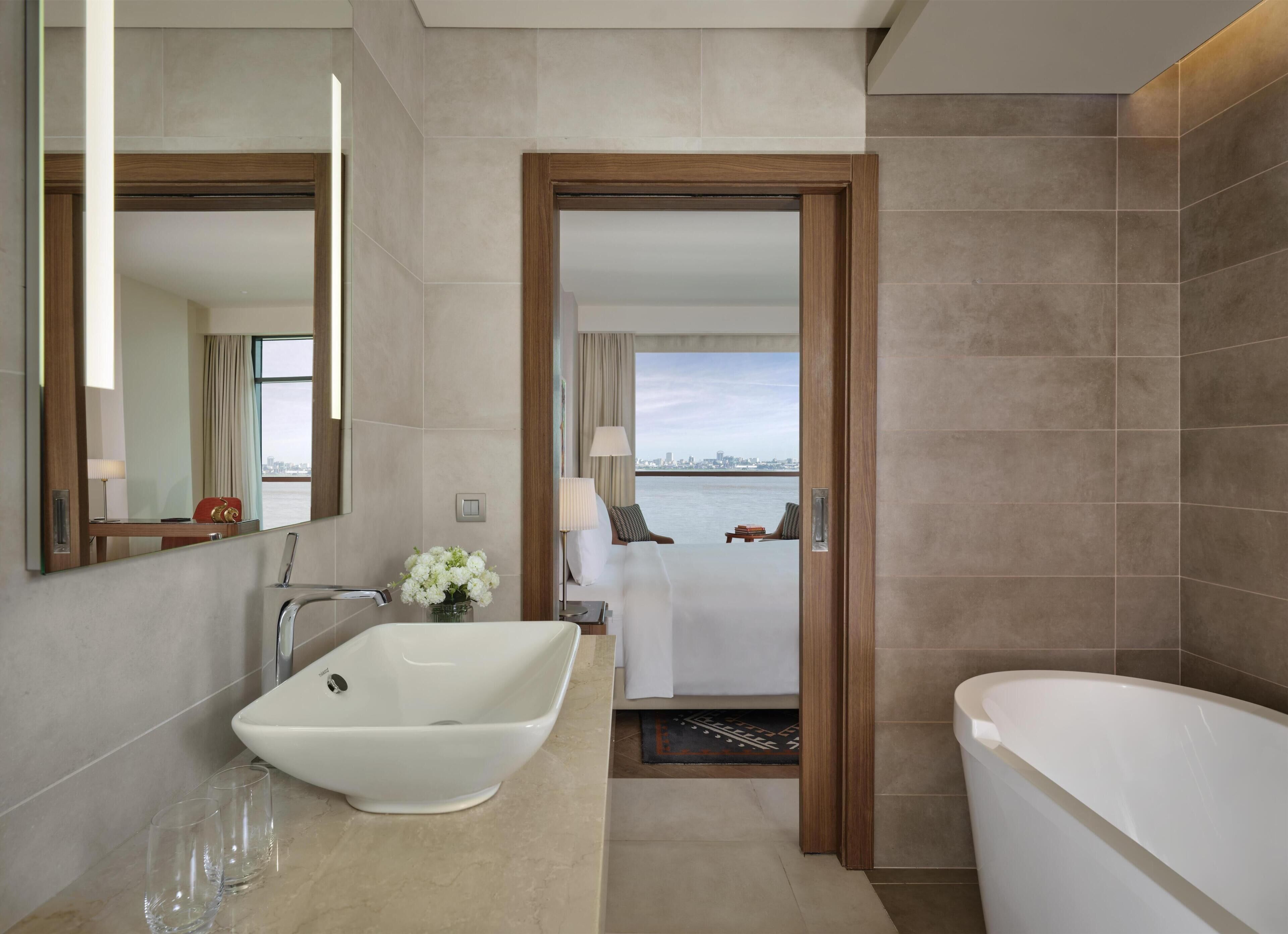 A modern bathroom with a freestanding bathtub, a large mirror, and a view of the city through an open door.