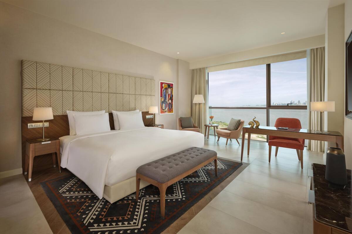 A hotel room with a large bed, a desk, a chair, a bench, and a view of the water.