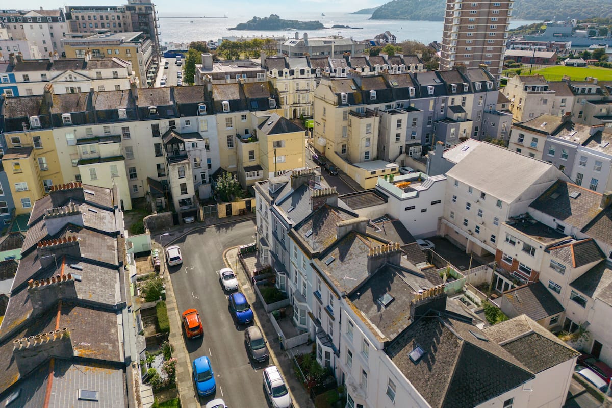 Il s’agit d’un quartier résidentiel où se côtoient des maisons mitoyennes et un grand immeuble d’appartements. Plusieurs voitures sont garées le long de la rue, et on aperçoit une étendue d’eau au loin.
