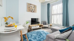 Uma sala de estar moderna com área de jantar, televisão de tela plana, sofá com almofadas, tapete azul e um quadro na parede.