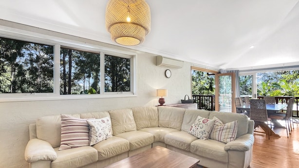 A living room with a large L-shaped sofa, a wooden coffee table, and a hanging light fixture. There are large windows with a view of trees and a balcony with outdoor furniture. - Riverside 3-bed Townhouse With Spacious Deck & BBQ (Mossy Point)